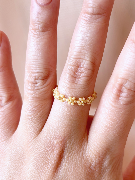 A close-up of a hand wearing a delicate daisy chain flower ring made of premium gold and neutral-toned Japanese seed beads.