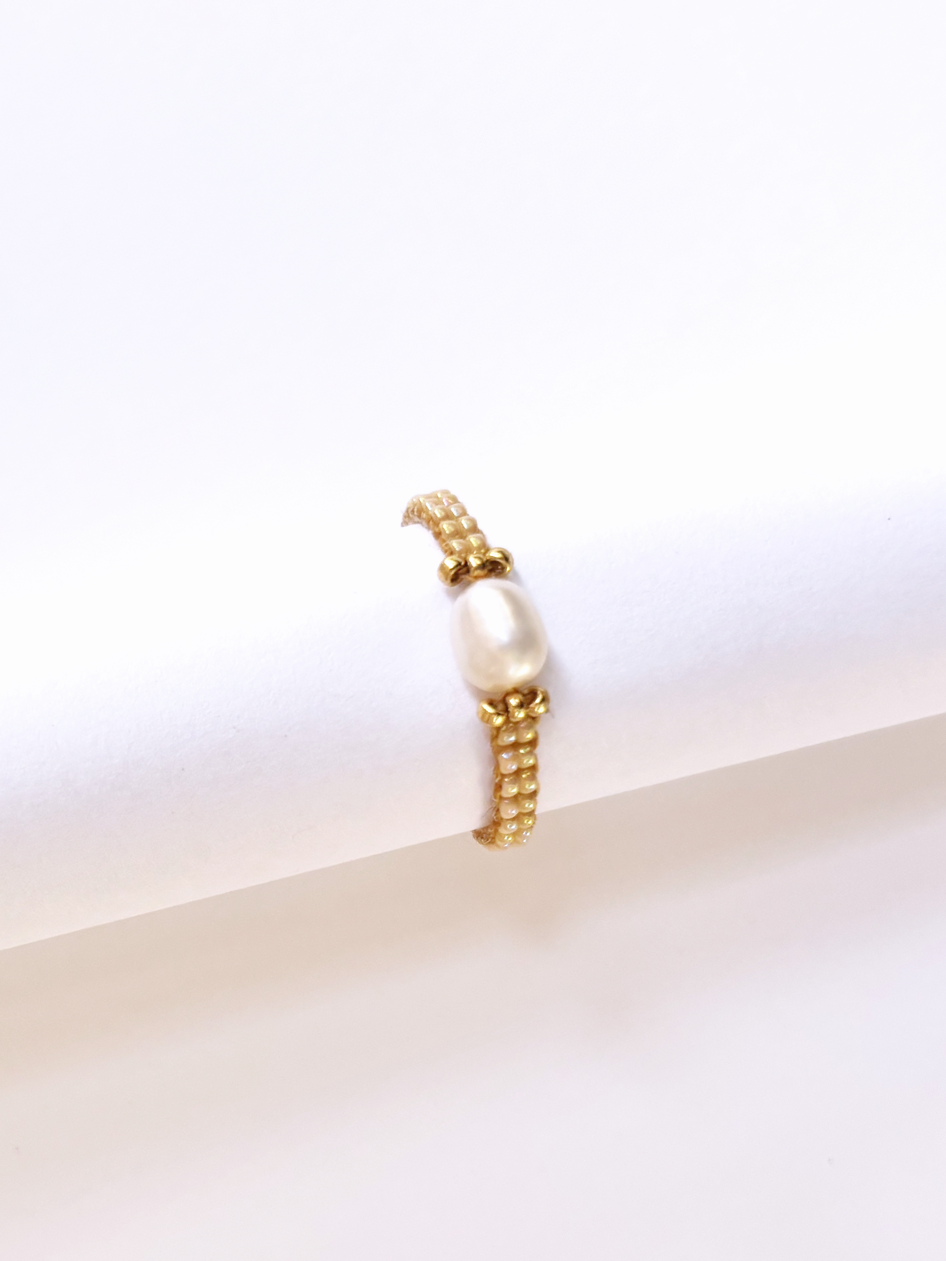 A delicate flower ring made of premium gold and neutral-toned Japanese seed beads with a single white pearl centerpiece, displayed on a jewelry stand.