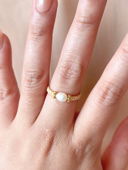 A close-up of a hand wearing a delicate ring made of premium Japanese seed beads in pearl and gold tones, featuring a central white pearl.