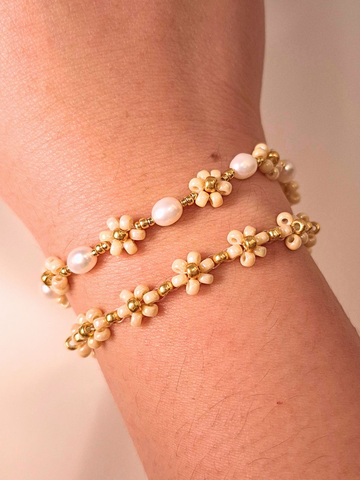 A close-up of a person's wrist styling a stacked set of two delicate handmade bracelets: the August Pearl Bracelet featuring genuine white freshwater pearls, and the minimalist cream Daisy Bracelet (August Breeze) with gold flower motifs, set against a soft beige fabric background.