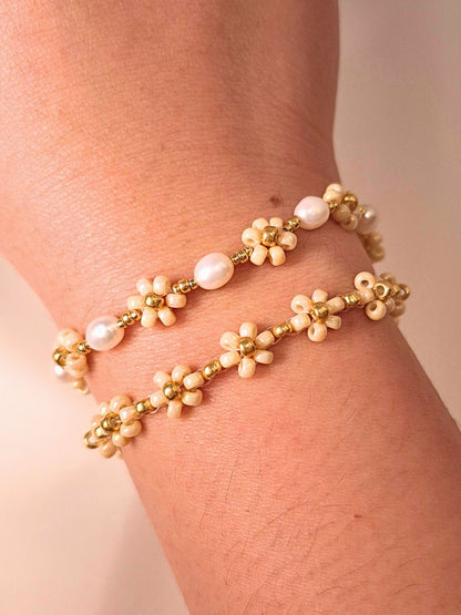 A close-up of a person's wrist styling a stacked set of two delicate handmade bracelets: the August Pearl Bracelet featuring genuine white freshwater pearls, and the minimalist cream Daisy Bracelet (August Breeze) with gold flower motifs, set against a soft beige fabric background.